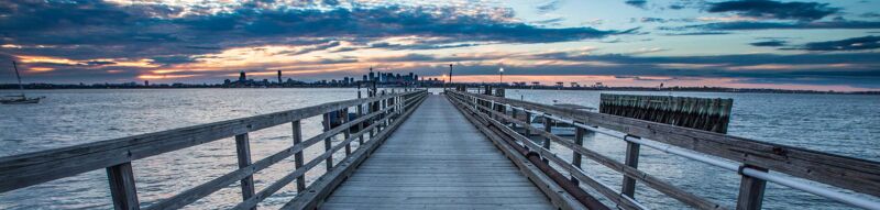 The image shows a long wooden pier extending into a body of water, likely a bay or ocean. The sky is filled with dramatic clouds, and the sun is setting, casting a warm glow on the horizon. In the distance, a city skyline is visible. The overall mood is peaceful and serene.